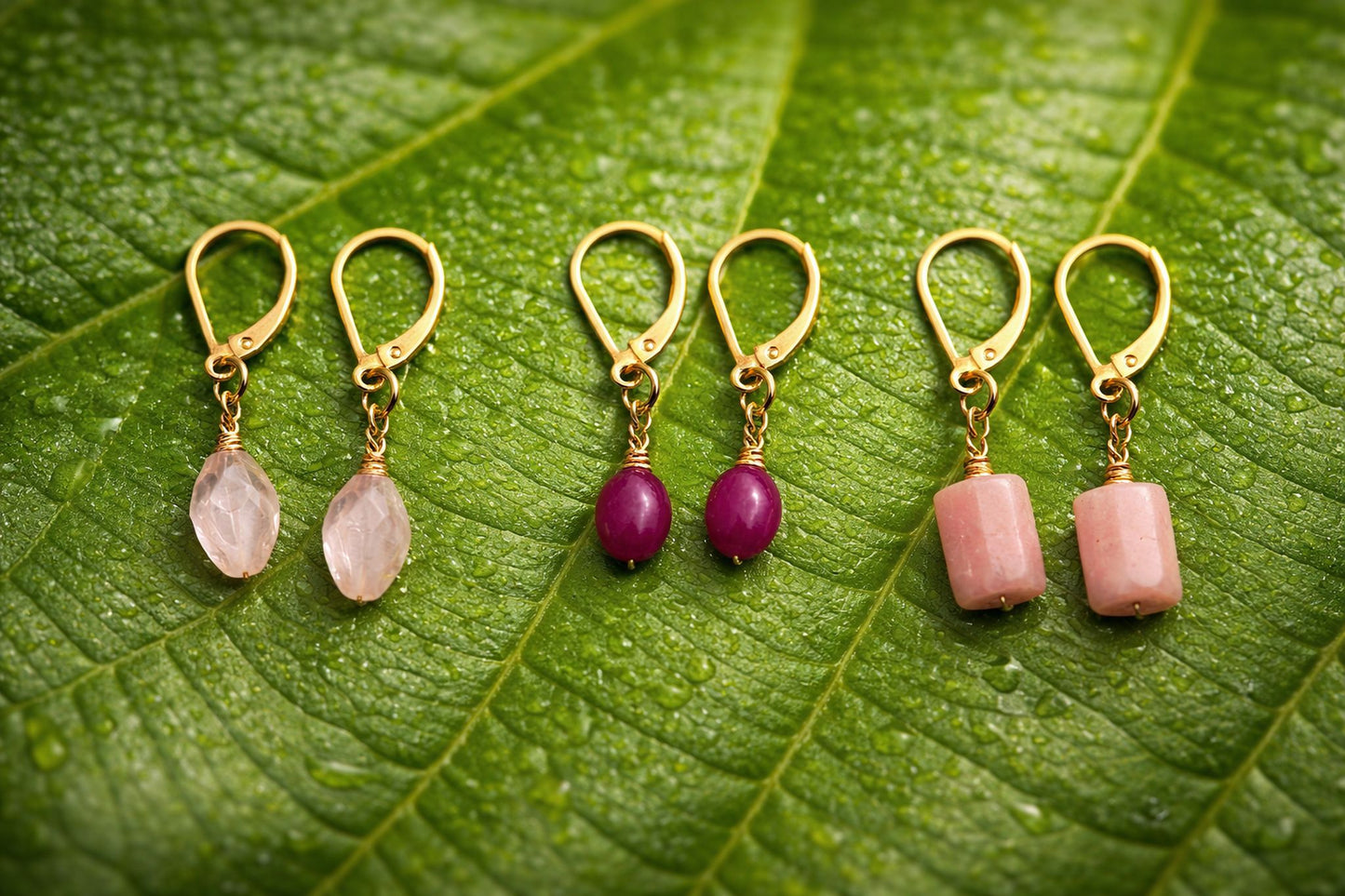 Set of three gold earrings with pink and purple gemstones on a green leaf background